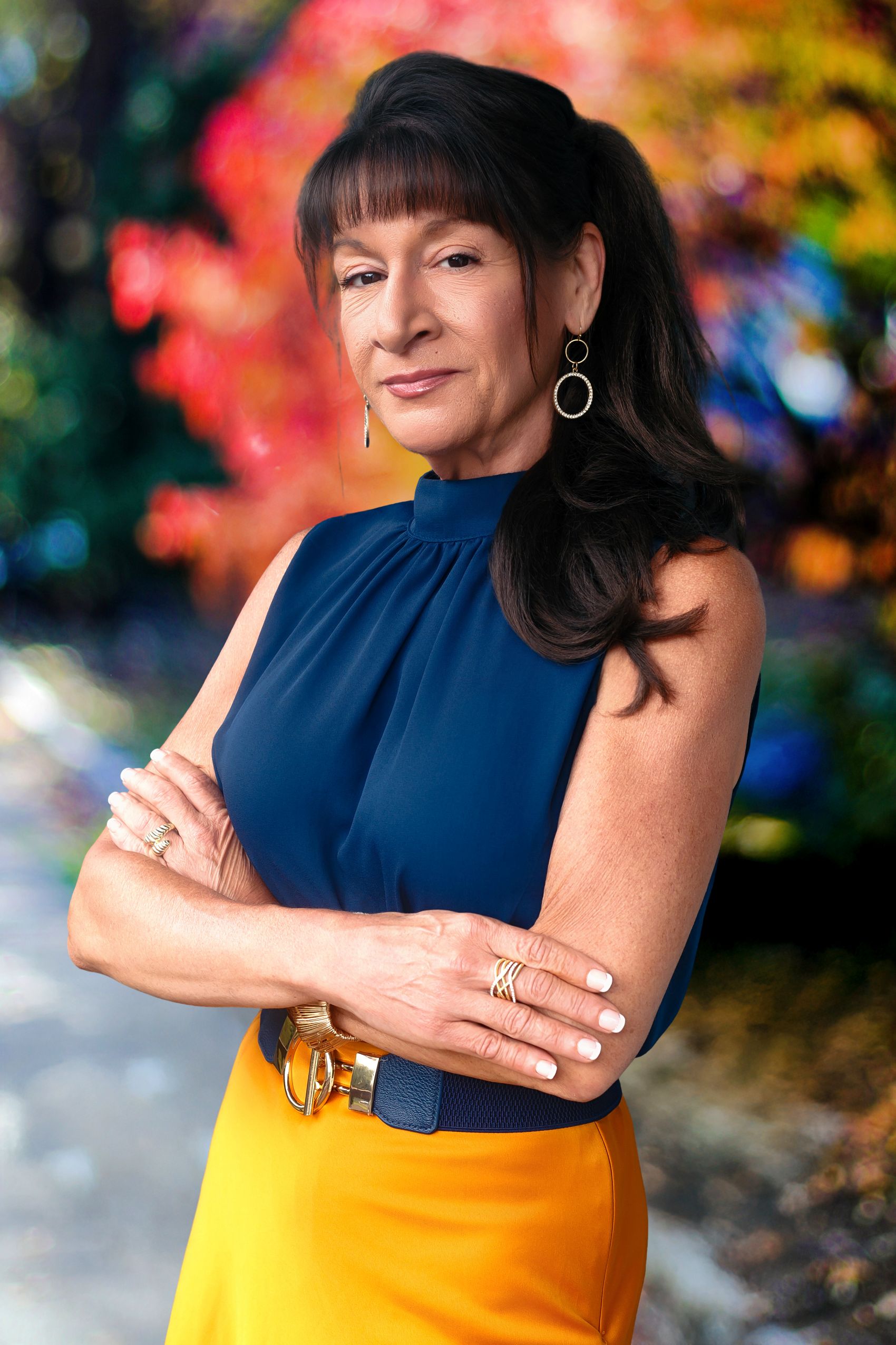 Gretchen Schneider headshot. Portrait of a confident woman with dark hair, wearing a blue sleeveless top and orange skirt, arms crossed in front of a colorful blurred background.